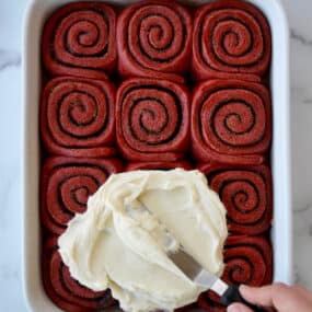 A white baking pan with red velvet cinnamon rolls and cream cheese frosting being slathered on top.