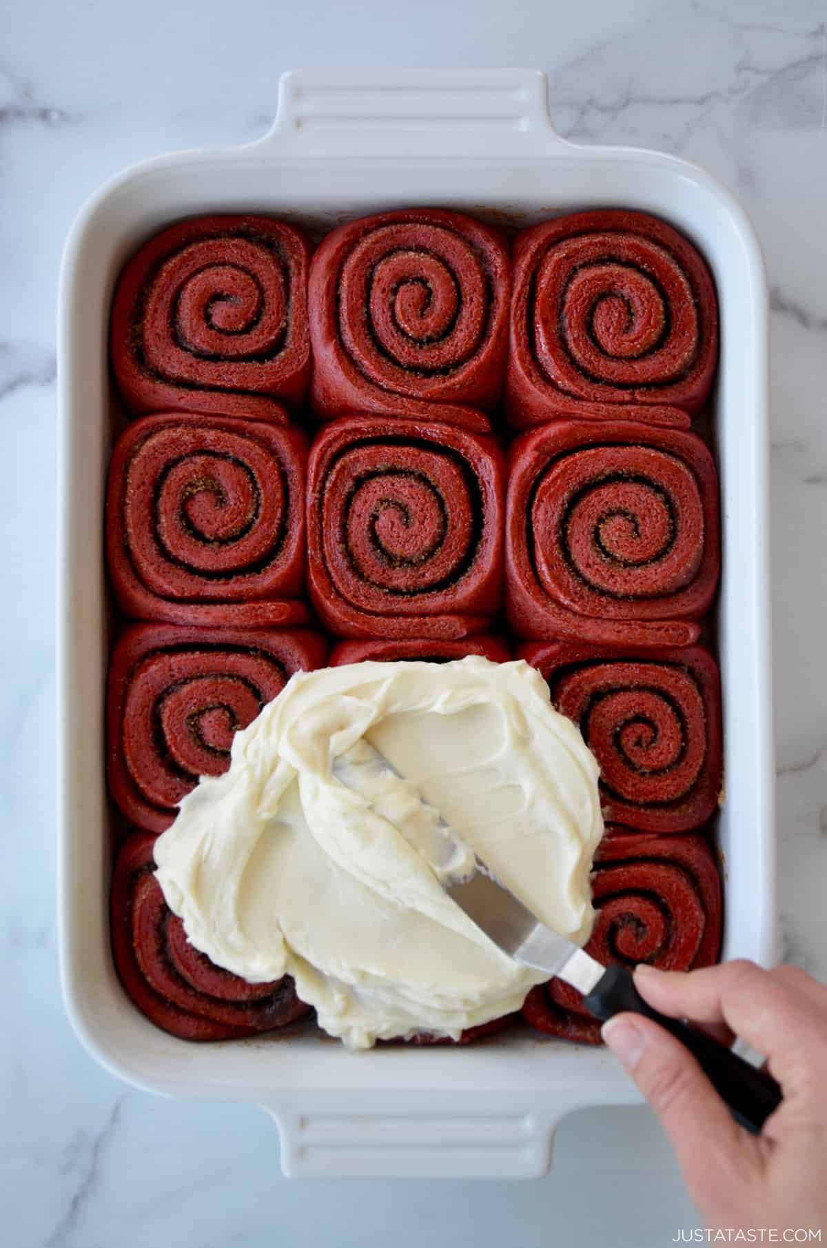 A white baking pan with red velvet cinnamon rolls and cream cheese frosting being slathered on top.