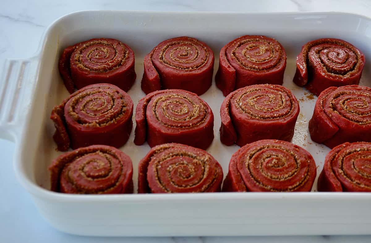 Uncooked red velvet cinnamon rolls are arranged in a white baking dish.