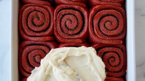 A white baking pan with red velvet cinnamon rolls and cream cheese frosting being slathered on top