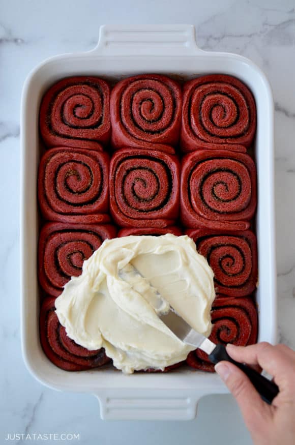 A white baking pan with red velvet cinnamon rolls and cream cheese frosting being slathered on top