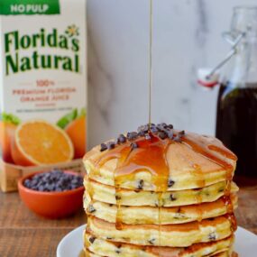 Stack of pancakes on a white plate with a carton of orange juice behind it