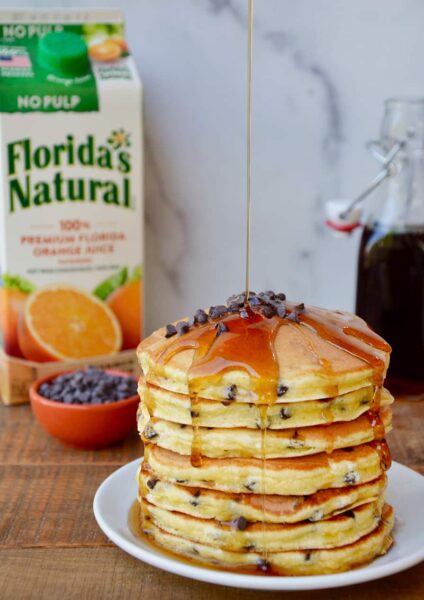 Stack of pancakes on a white plate with a carton of orange juice behind it