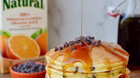 Stack of pancakes on a white plate with a carton of orange juice behind it