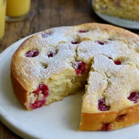 A thick coffee cake studded with raspberries with a slice missing on a round platter next to two glasses filled with orange juice.