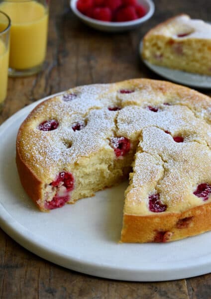 A thick coffee cake studded with raspberries with a slice missing on a round platter next to two glasses filled with orange juice.