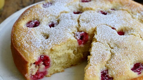 A thick coffee cake studded with raspberries with a slice missing on a round platter next to two glasses filled with orange juice.