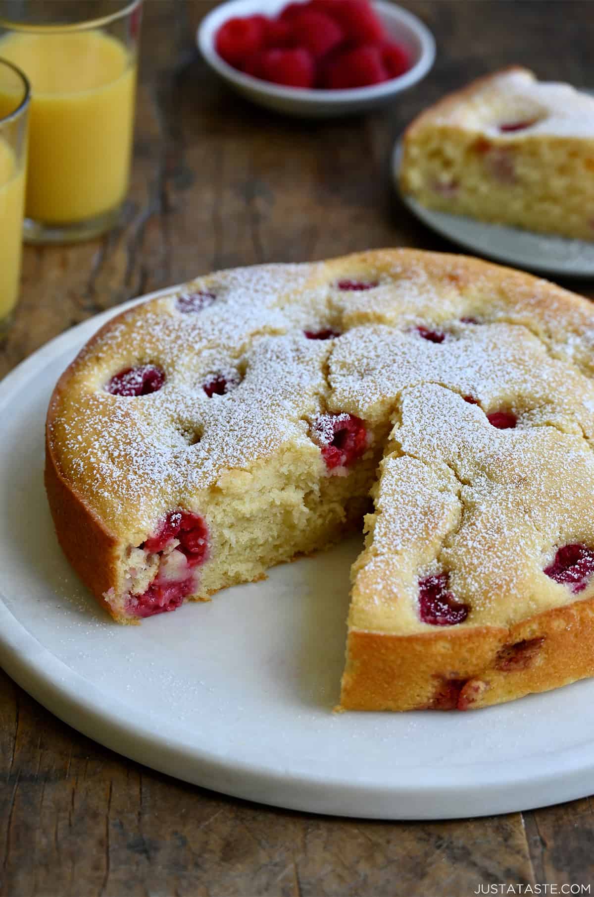 A thick coffee cake studded with raspberries with a slice missing on a round platter next to two glasses filled with orange juice.