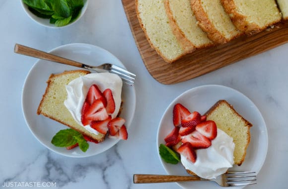 Two dessert plates with a slice of Cream Cheese Pound Cake topped with whipped cream, fresh mint leaves and sliced strawberries next to cutting board with more slices of cake