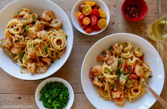 Two bowls containing the best Quick Shrimp Scampi next to a glass of wine and small bowls containing sliced cherry tomatoes, chili flakes and chopped parsley