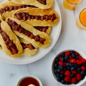 White plate containing Bacon Pancake Dippers next to glasses of orange juice, small bowl with berries and hand dunking pancake dipper in maple syrup