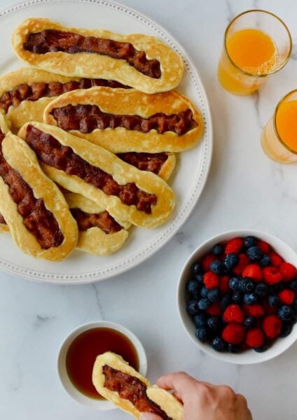White plate containing Bacon Pancake Dippers next to glasses of orange juice, small bowl with berries and hand dunking pancake dipper in maple syrup