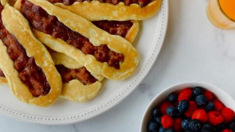 White plate containing Bacon Pancake Dippers next to glasses of orange juice, small bowl with berries and hand dunking pancake dipper in maple syrup