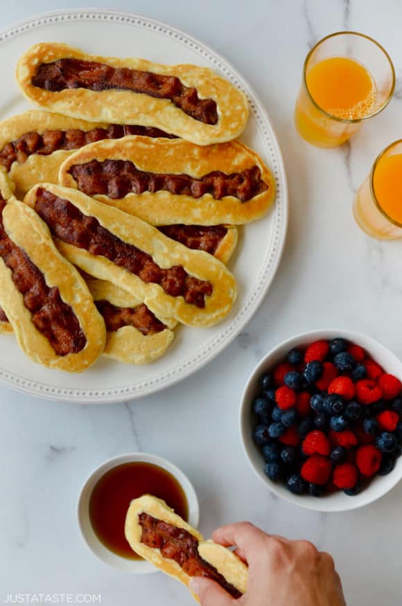 White plate containing Bacon Pancake Dippers next to glasses of orange juice, small bowl with berries and hand dunking pancake dipper in maple syrup