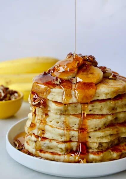 A tall stack of banana nut pancakes topped with slices of banana, chopped pecans and maple syrup.