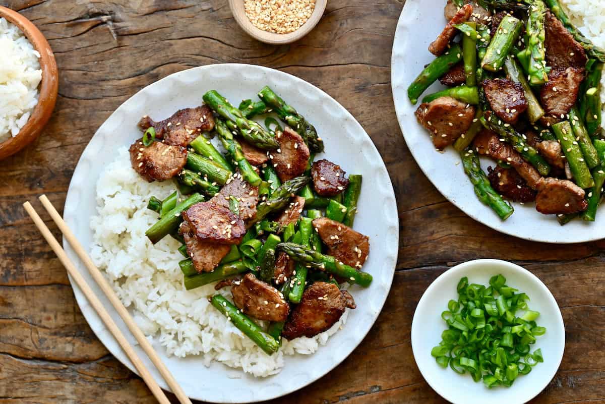 Pieces of black pepper pork and asparagus on a dinner plate with steamed white rice and chopsticks.