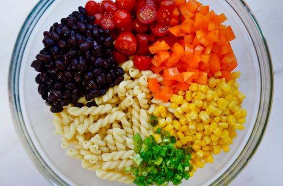 Glass bowl containing diced tomatoes, diced orange bell pepper, yellow corn kernels, sliced scallions, cavatappi and black beans 