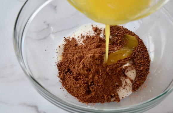 A glass bowl filled with brownie batter ingredients and butter being poured in