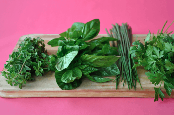A wood cutting board with fresh herbs on top and a pink background