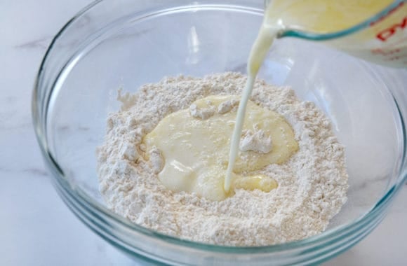 A large glass bowl containing flour with buttermilk and melted butter being poured in