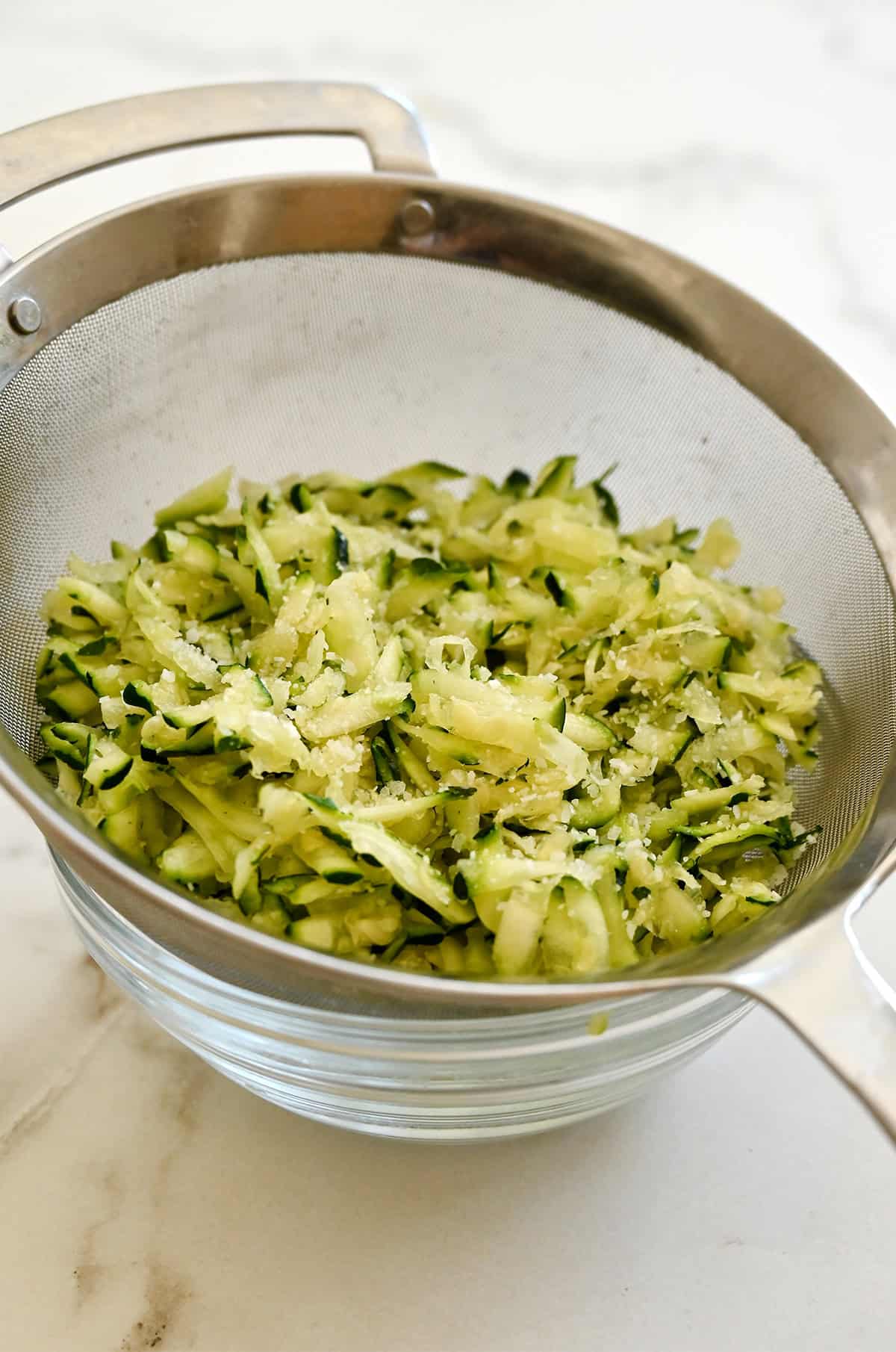 Salted shredded zucchini in a sieve, draining over a glass bowl.