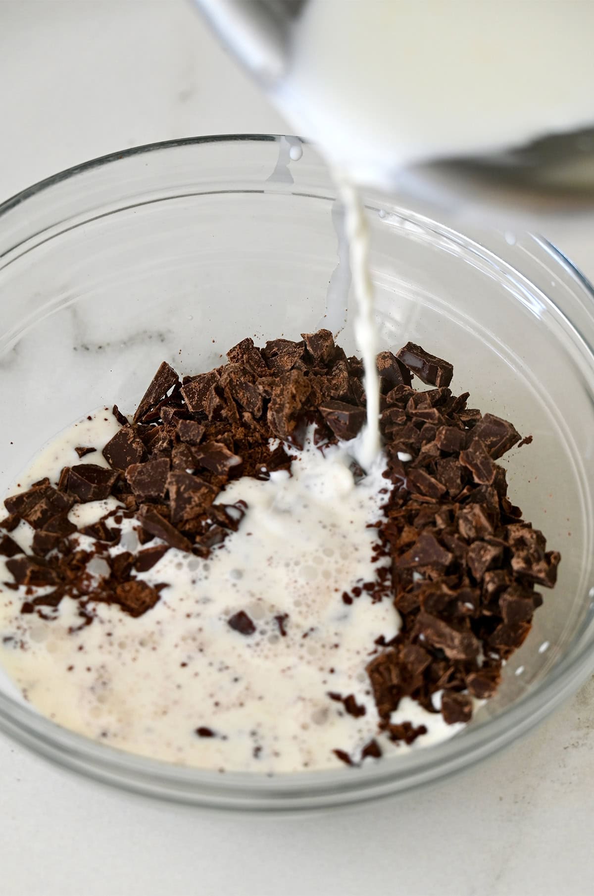 Warmed heavy cream being poured over chopped chocolate in a glass bowl.