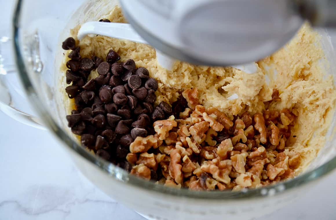 Bowl of a stand mixer containing cookie dough mixture, walnuts and chocolate chips