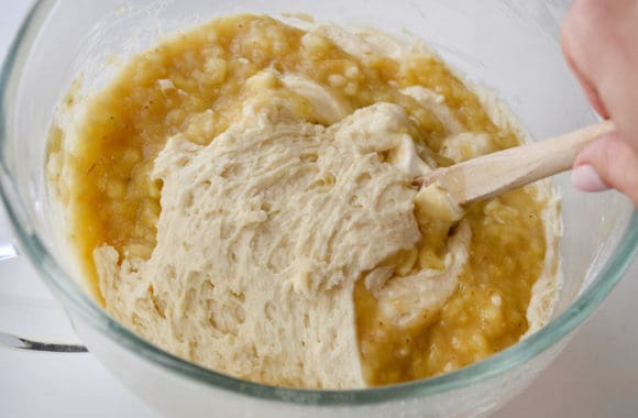 Hand holding spatula in bowl containing mashed bananas and batter