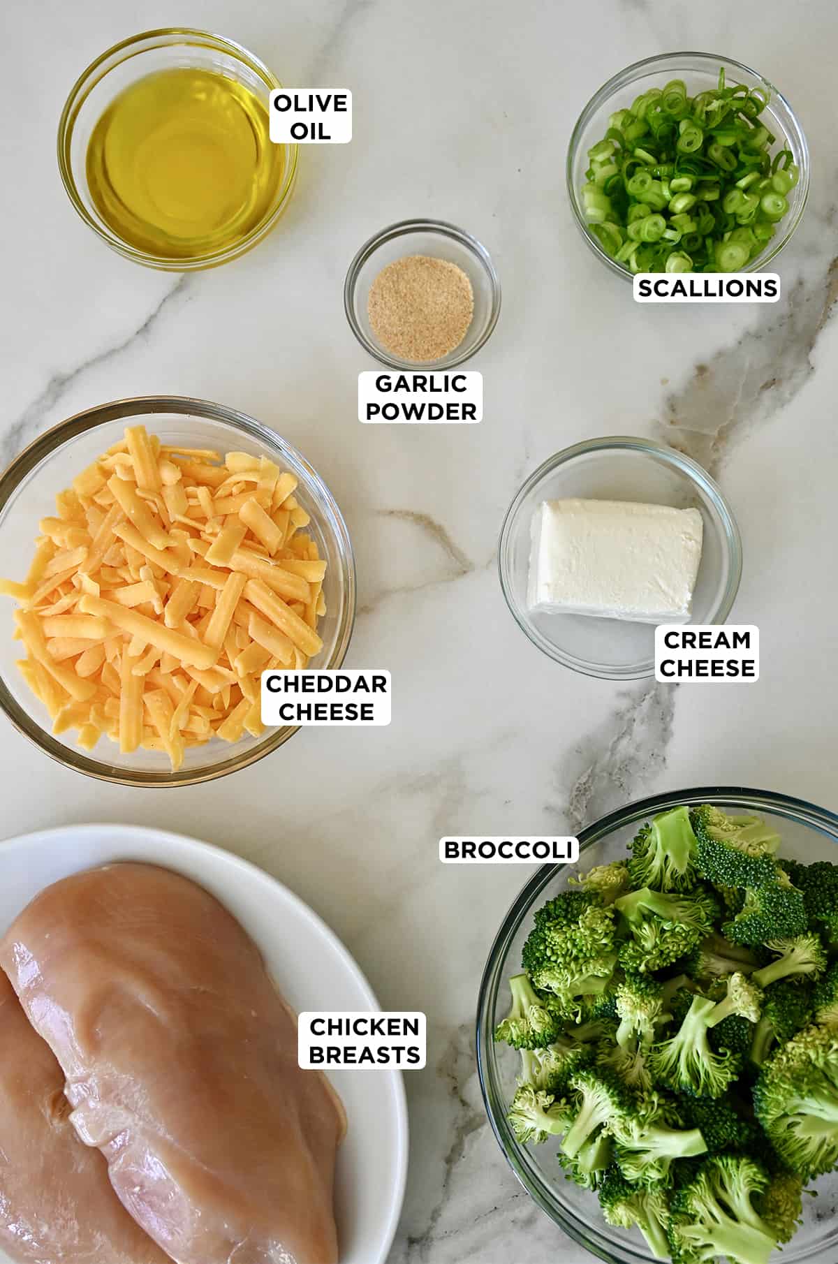 Various sizes of glass bowls containing stuffed chicken ingredients, including olive oil, garlic powder, scallions, cream cheese, cheddar cheese, broccoli and chicken breasts.