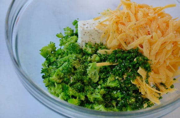A glass bowl containing chopped broccoli, shredded cheddar cheese and cream cheese