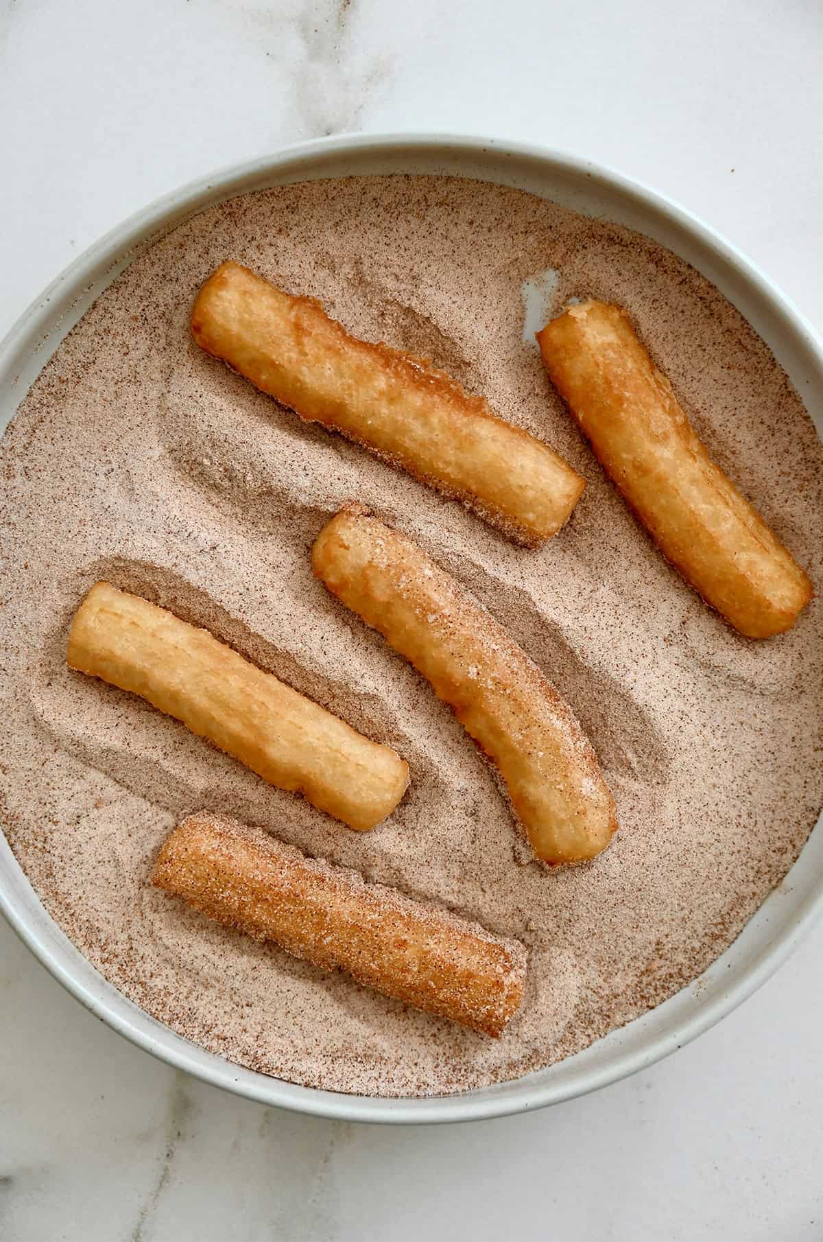 Warm churros are being rolled in cinnamon sugar on a shallow plate.