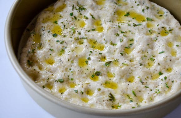 Round baking pan with focaccia dough drizzled with olive oil and fresh herbs