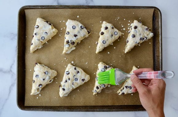 Hand with brush brushing tops of chocolate chip scones with heavy cream 