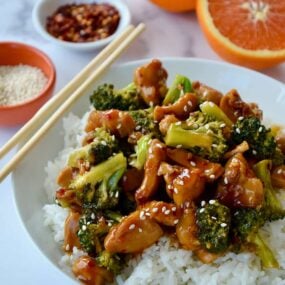 Quick Orange Chicken and Broccoli over white rice in bowl with chopsticks next to a sliced orange