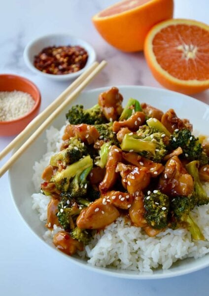 Quick Orange Chicken and Broccoli over white rice in bowl with chopsticks next to a sliced orange