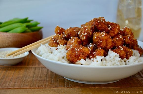 A white bowl with sesame chicken on top of white rice with a bowl of edamame in the background