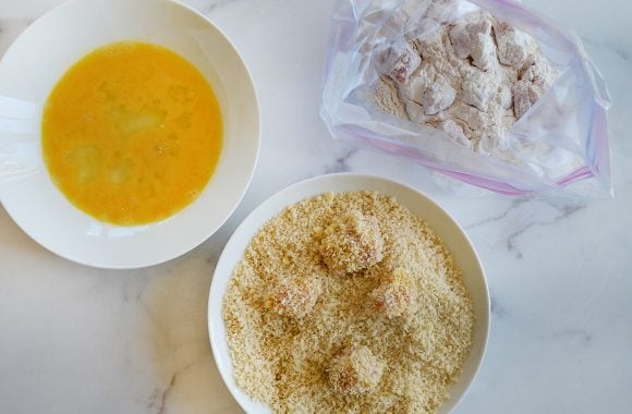 Bowls containing beaten eggs and breadcrumbs and a bag with chicken nuggets in flour
