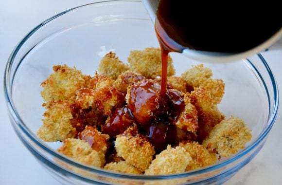 A glass bowl containing crispy chicken thigh pieces with sesame sauce being poured on top