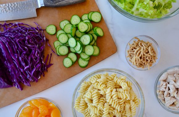 Cutting board with shredded red cabbage, diced cucumbers and knife next to small bowls containing lettuce, chow mein noodles, shredded chicken, pasta and mandarin oranges