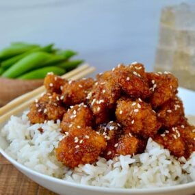 A white bowl containing white rice topped with sesame chicken and chopsticks on the side