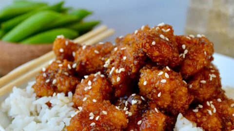 A white bowl containing white rice topped with sesame chicken and chopsticks on the side