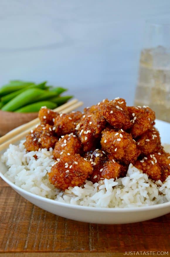 A white bowl containing white rice topped with sesame chicken and chopsticks on the side