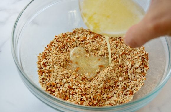 Melted butter being poured over crushed pretzels in clear glass bowl