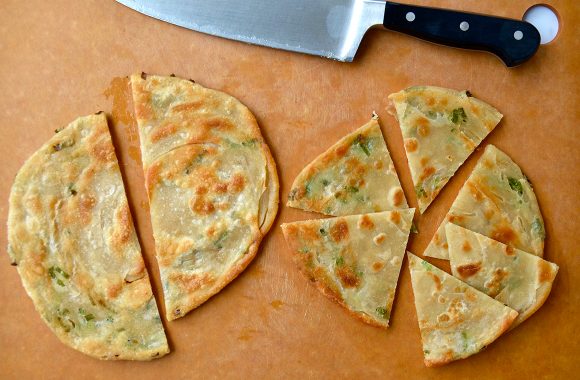 A cutting board with a knife and scallion pancakes cut into wedges