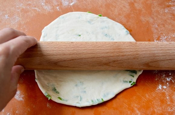 A rolling pin on top of a scallion pancake on a cutting board