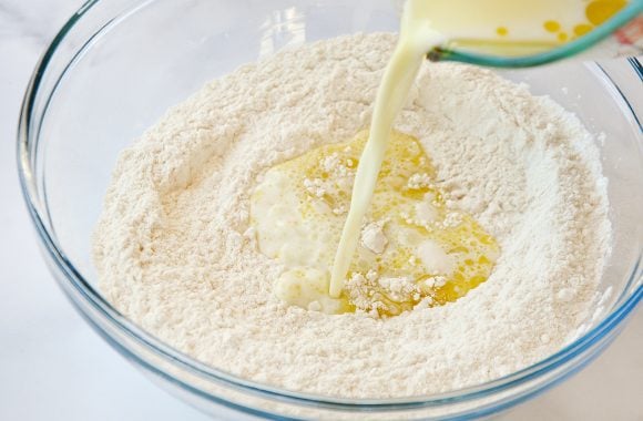 Milk and olive oil being poured from measuring cup into bowl containing flour 