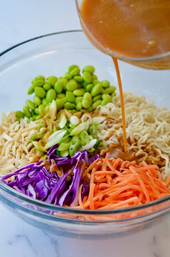 Peanut dressing being poured over carrots, cabbage, scallions, edamame and noodles in a bowl