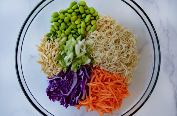 Clear bowl containing ramen noodles, shredded carrots and purple cabbage, sliced scallions, slivered almonds and shelled edamame