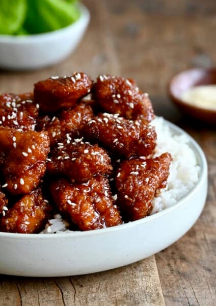 Crispy sesame chicken topped with sesame seeds over white rice on a dinner plate.