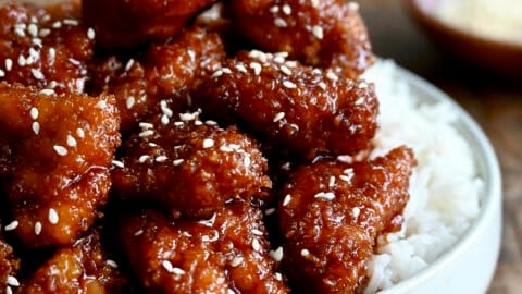 Crispy sesame chicken topped with sesame seeds over white rice on a dinner plate.